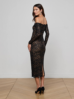 A woman stands on a wooden floor, wearing the Lindsay Sequined Lace Dress by L’AGENCE—an off-shoulder black lace gown with long sleeves, side slit, and black heels. She looks over her shoulder against a plain light background.
