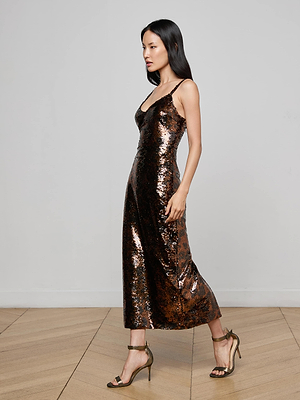 A woman in the L'AGENCE Noella Sequin Dress—a sleeveless, bronze, chic midi—pairs it with high-heeled sandals as she walks indoors on a light wooden floor against a plain white wall.