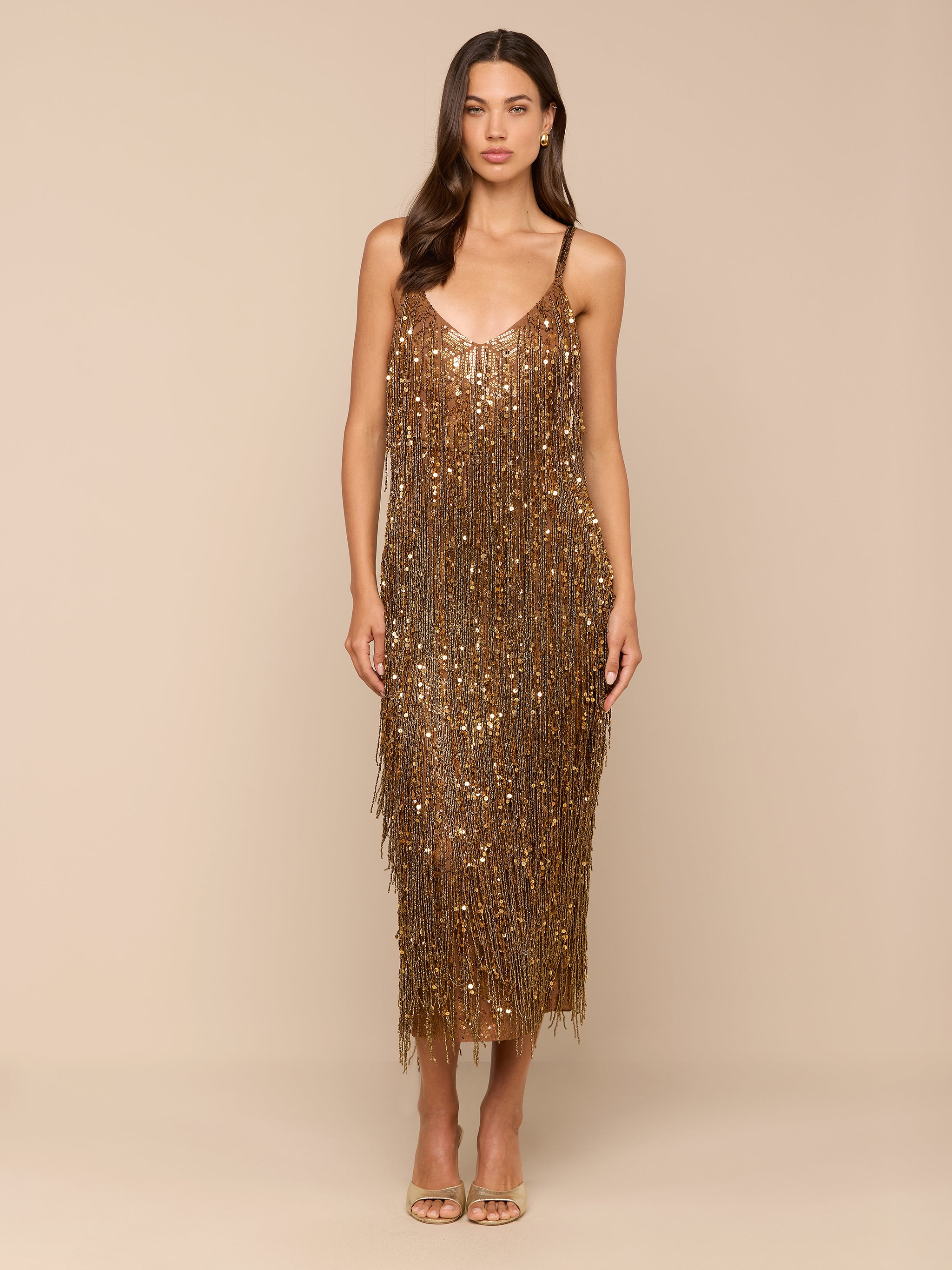 A woman stands against a plain background, wearing the Noella Sequin Fringe Dress—an elegant gold sequin dress with fringe embellishments and thin straps—paired with open-toe heels. Her long, wavy brown hair frames her face, which holds a neutral expression.