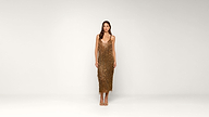 A woman stands against a plain white background wearing a gold, sequined, sleeveless dress with thin straps and gold open-toe heels.