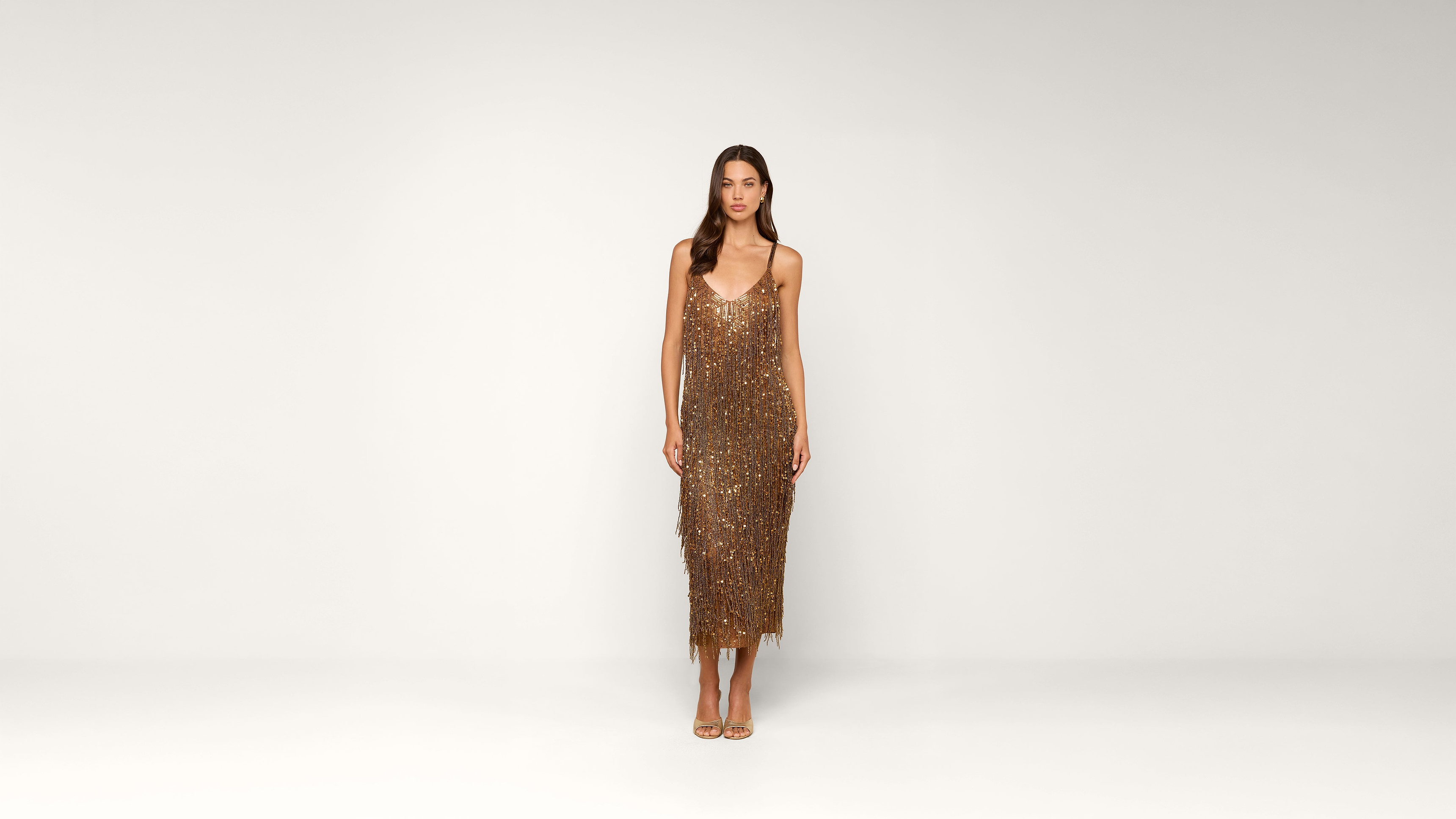 A woman stands against a plain white background wearing a gold, sequined, sleeveless dress with thin straps and gold open-toe heels.