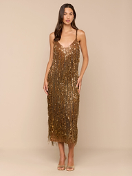 A woman stands facing forward, modeling the Noella Sequin Fringe Dress—a midi-length gold sequin dress with fringe embellishments and thin straps. She has long, loose hair and wears nude heels against a plain white background.