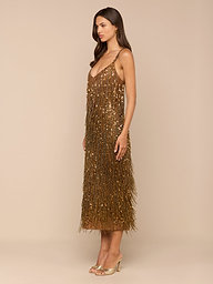 A woman stands sideways against a plain white background, wearing the Noella Sequin Fringe Dress—a gold, midi-length design adorned with sequin and fringe embellishments—paired with open-toe heels.
