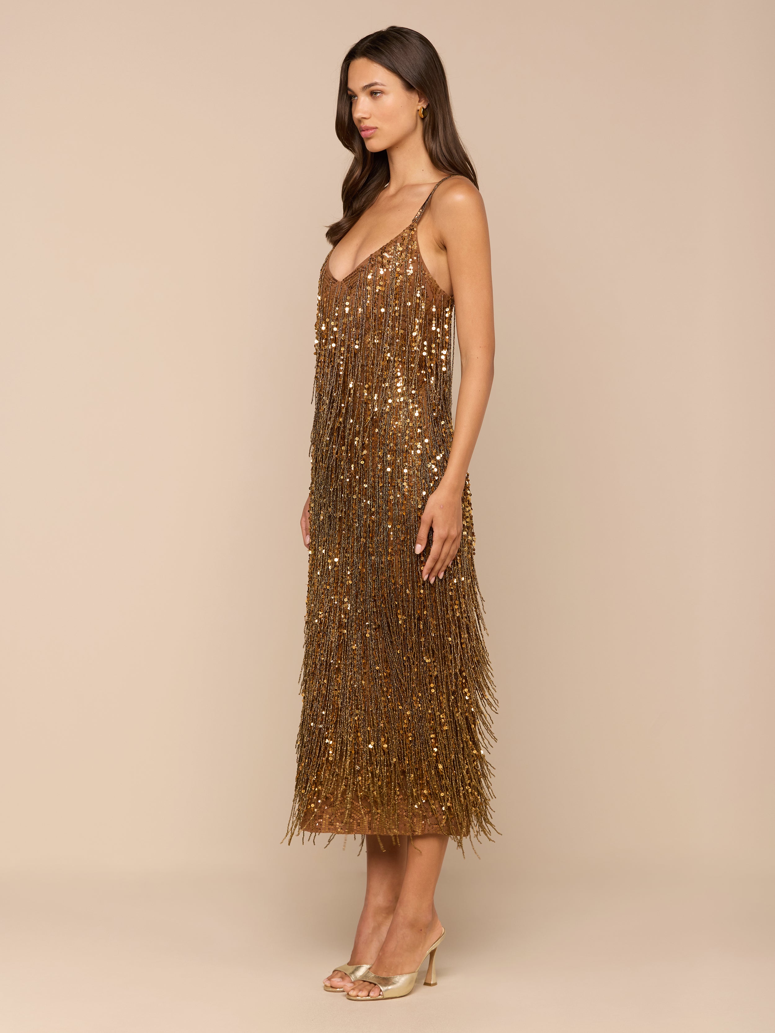 A woman stands sideways against a plain white background, wearing the Noella Sequin Fringe Dress—a gold, midi-length design adorned with sequin and fringe embellishments—paired with open-toe heels.