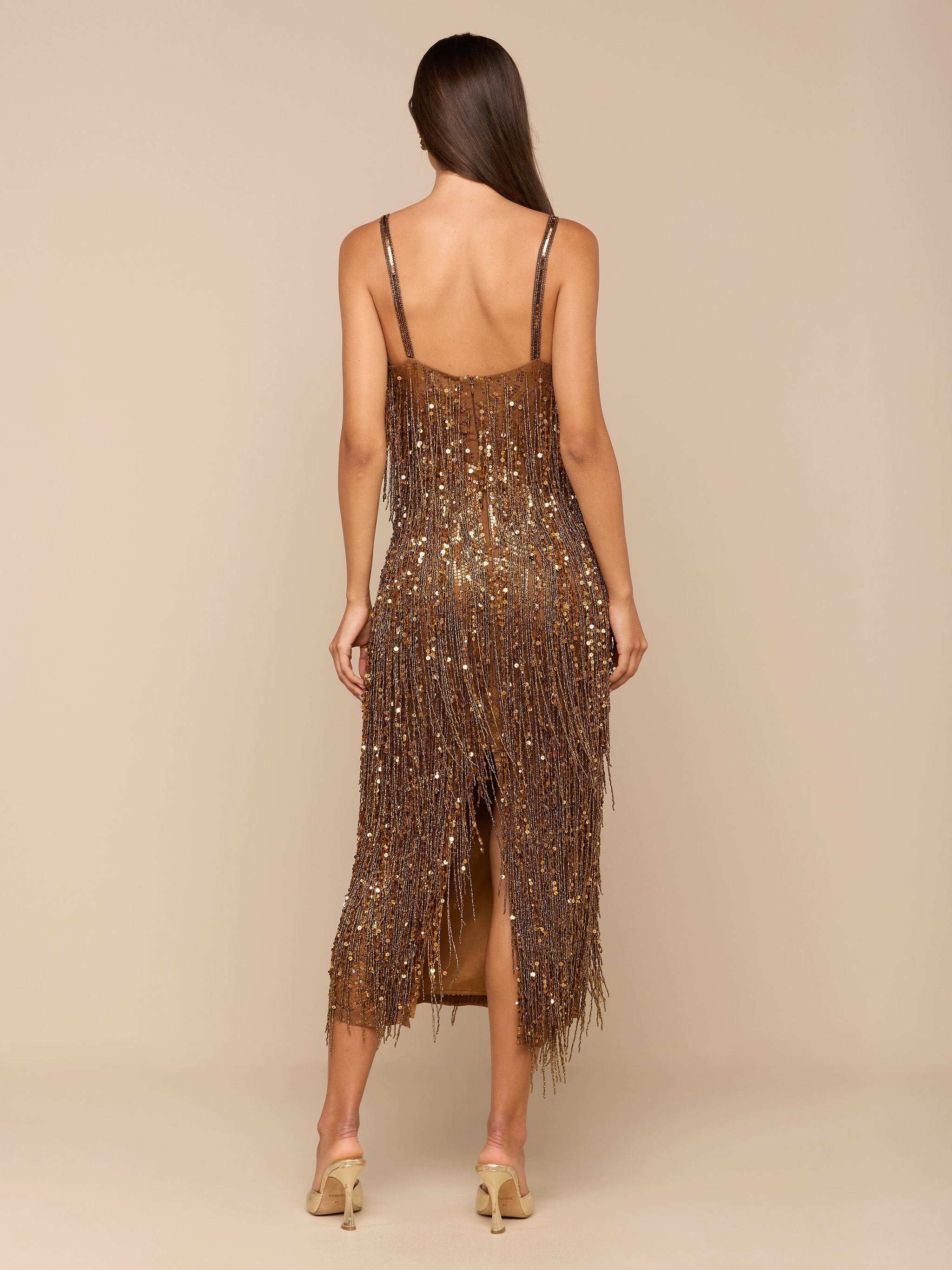 A woman stands with her back to the camera against a plain, light background, wearing the Noella Sequin Fringe Dress—a gold, midi-length dress adorned with shimmering sequins and fringe embellishments—paired with strappy heels.