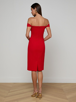 A woman faces away, wearing the Vivie Dress—a red piece with off-the-shoulder straps and a sculpted corset bodice. She pairs it with gold heels and earrings, her straight brown hair contrasting a light wall and wood-patterned floor.