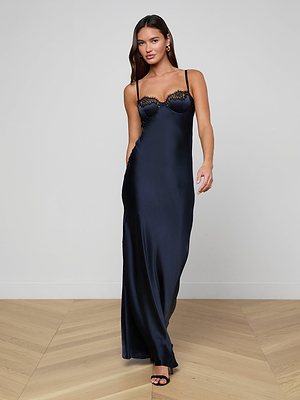 A woman walks on a light wooden floor against a white wall, wearing the Shayla Silk Lace Gown—an event-ready dark blue gown featuring lace detailing on the bodice.
