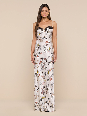 A woman with long brown hair and a neutral expression stands against a beige background, wearing the Sayla Silk Gown—a long, sleeveless white silk dress featuring butterfly lock print and black lace trim at the bodice.
