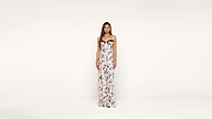 A woman stands against a plain white background, wearing a long, white, strapless dress with a colorful floral pattern.