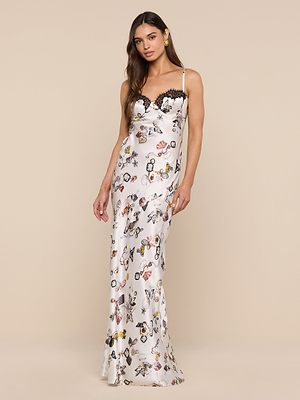 A woman in the Sayla Silk Gown—a sleeveless, floor-length white satin dress with a vibrant butterfly lock print and black lace neckline—stands against a plain beige background.