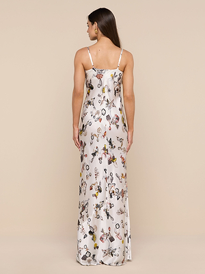 A woman faces away in the Sayla Silk Gown—a long white dress with thin straps and a vibrant butterfly lock print—set against a plain beige background.