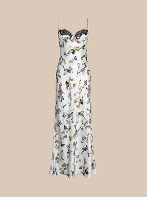 The Sayla Silk Gown is a white, sleeveless satin silk dress with thin straps, a black lace-trimmed sweetheart neckline, and a colorful butterfly lock print with illustrated figures and objects. Displayed on a plain beige background.