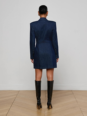 A person stands indoors with their back to the camera, wearing the L'AGENCE Kylan Blazer Dress in dark blue and black knee-high heeled boots on a light wooden floor with a plain white wall background.