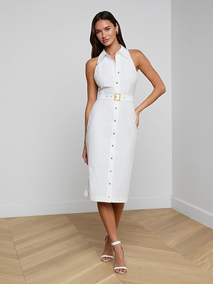 A woman stands indoors on wooden flooring wearing the sleeveless Lisette Midi Dress in organic cotton, featuring a belted waist, paired with white heeled sandals. She poses with one hand on her hip and a slight smile.