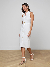 A woman walks on a wooden floor wearing the Lisette Midi Dress—sleeveless, button-down, organic cotton, with a belted waist and gold buckle—styled with white heeled sandals and hoop earrings.
