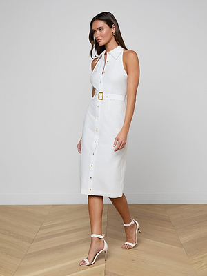 A woman walks on a wooden floor wearing the Lisette Midi Dress—sleeveless, button-down, organic cotton, with a belted waist and gold buckle—styled with white heeled sandals and hoop earrings.