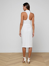 A woman stands on a wooden floor facing away from the camera, wearing the sleeveless Lisette Midi Dress in white organic cotton with a racerback design and white high-heeled shoes. The background is a plain white wall.