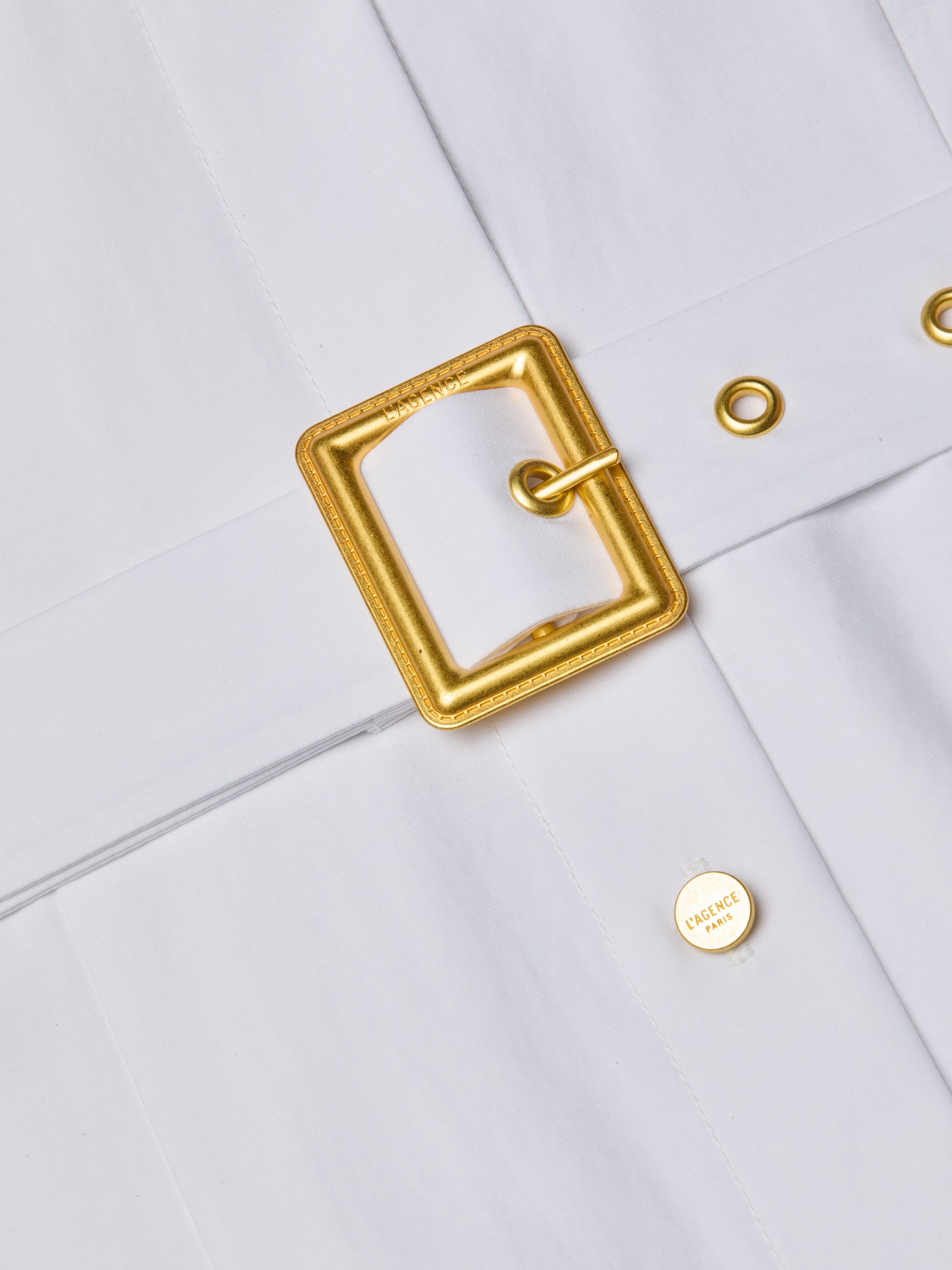 Close-up of a white organic cotton belt with a square gold buckle and gold eyelets, plus a small round gold HERMÈS button—ideal for pairing with your favorite racerback style or the Lisette Midi Dress.