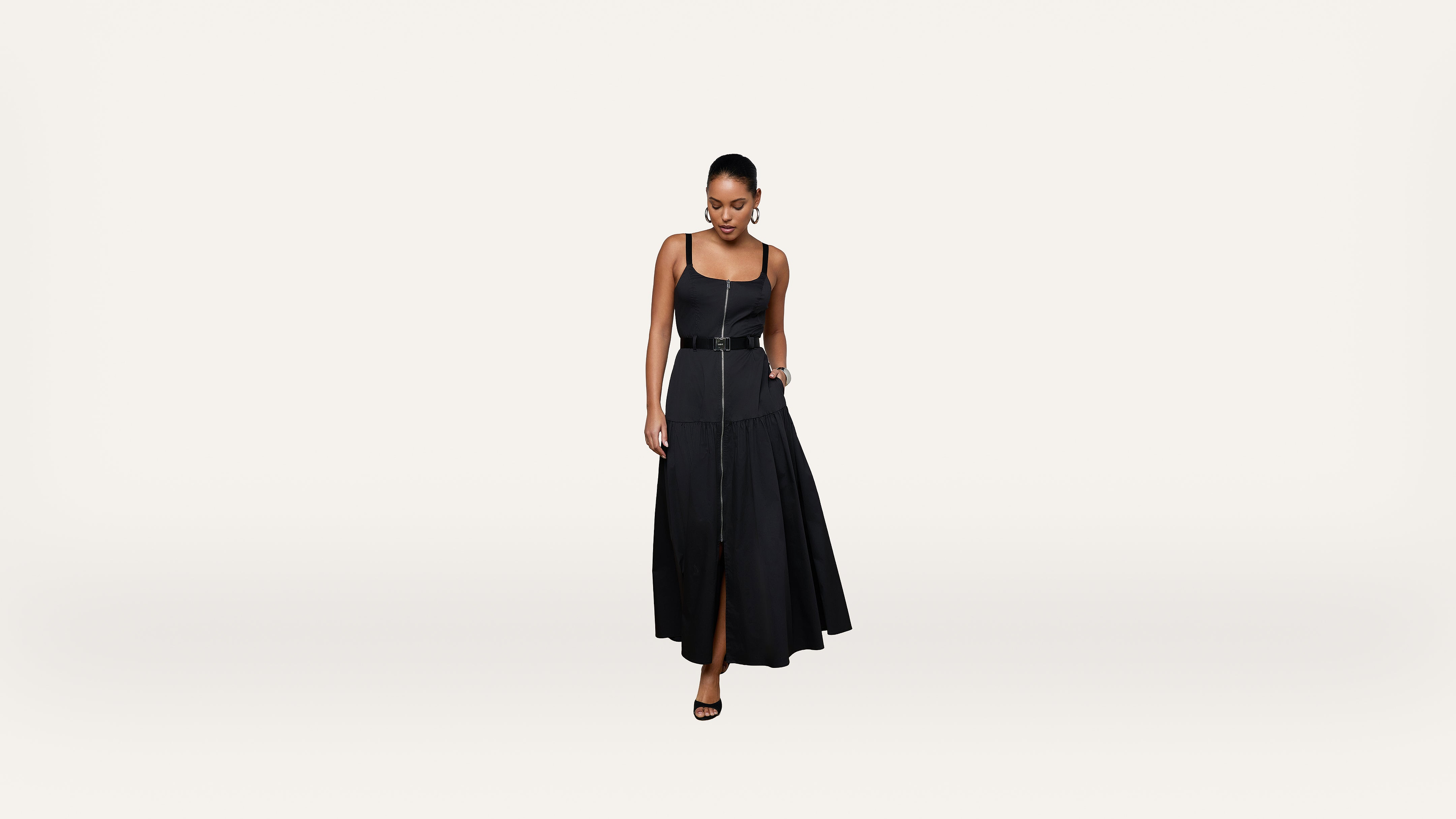 A woman wearing a sleeveless black dress with a belt and black heels stands against a plain white background, looking down with one hand holding her dress.