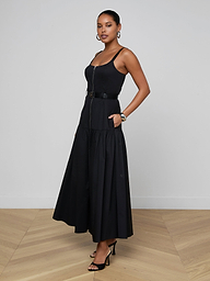 Wearing the Rima Maxi Dress—a sleeveless black stretch poplin dress featuring a belted waist and flared skirt—a woman stands with one hand in her pocket against a plain white background, styled with black heels and hoop earrings.