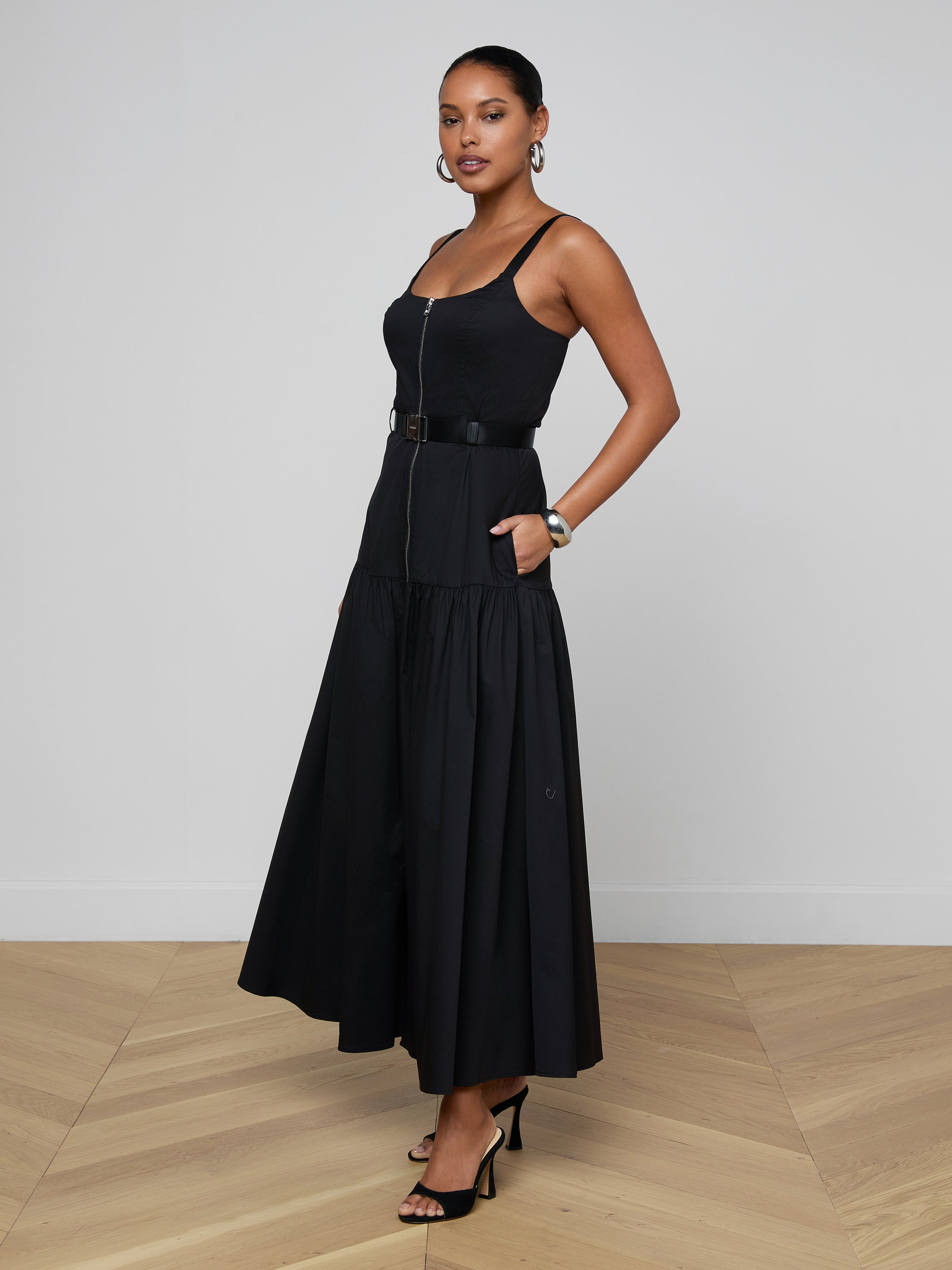 Wearing the Rima Maxi Dress—a sleeveless black stretch poplin dress featuring a belted waist and flared skirt—a woman stands with one hand in her pocket against a plain white background, styled with black heels and hoop earrings.