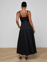 A woman stands facing away, wearing the Rima Maxi Dress—a sleeveless black stretch poplin dress featuring a fitted bodice and flared skirt—paired with black heels. The background is plain and light-colored.