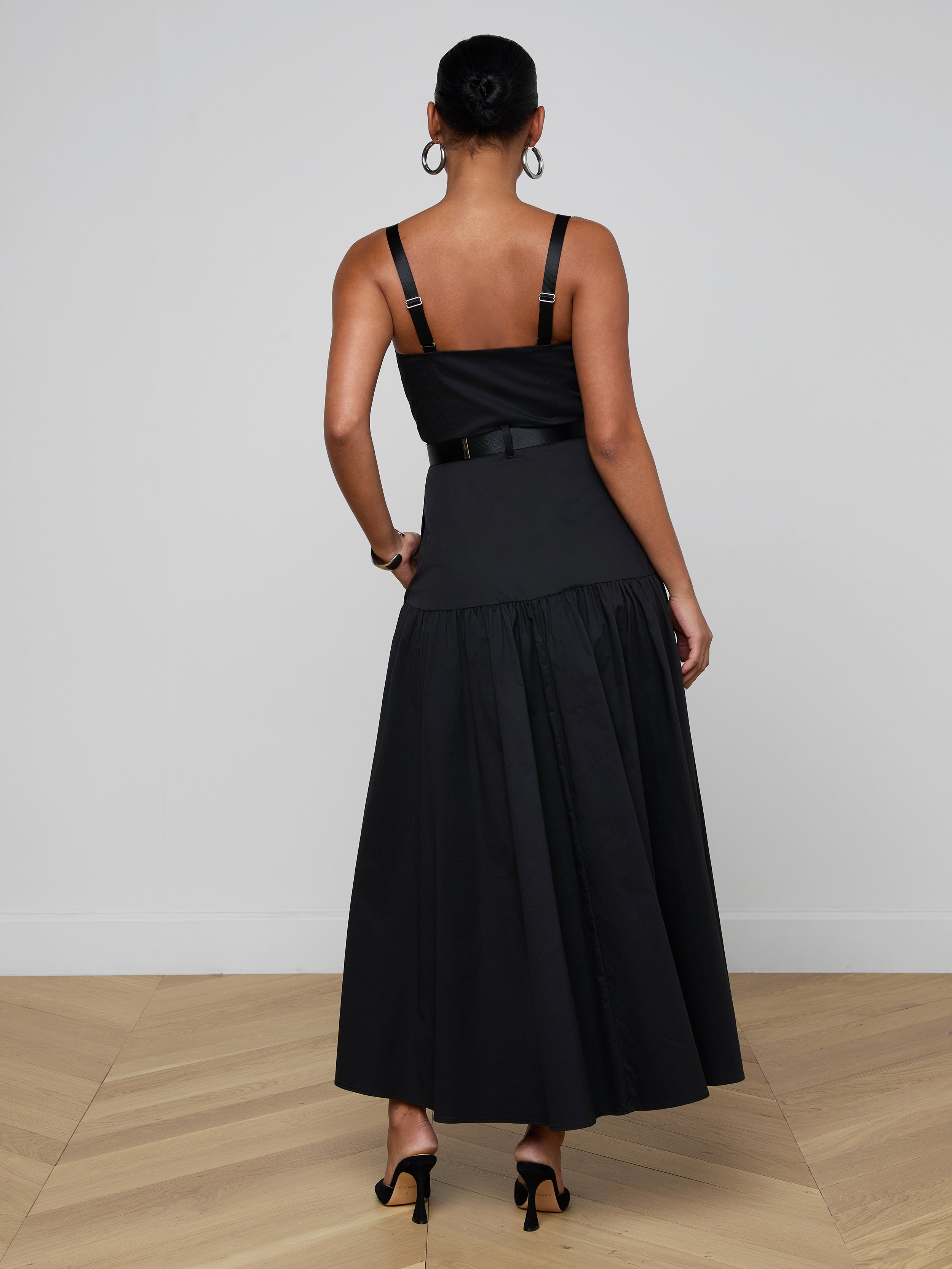 A woman stands facing away, wearing the Rima Maxi Dress—a sleeveless black stretch poplin dress featuring a fitted bodice and flared skirt—paired with black heels. The background is plain and light-colored.
