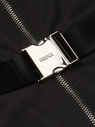 A close-up of a shiny silver buckle engraved with LAGENCE, fastened to a black strap on the Rima Maxi Dress in stretch poplin. Behind it, a metallic zipper runs vertically down the black fabric.