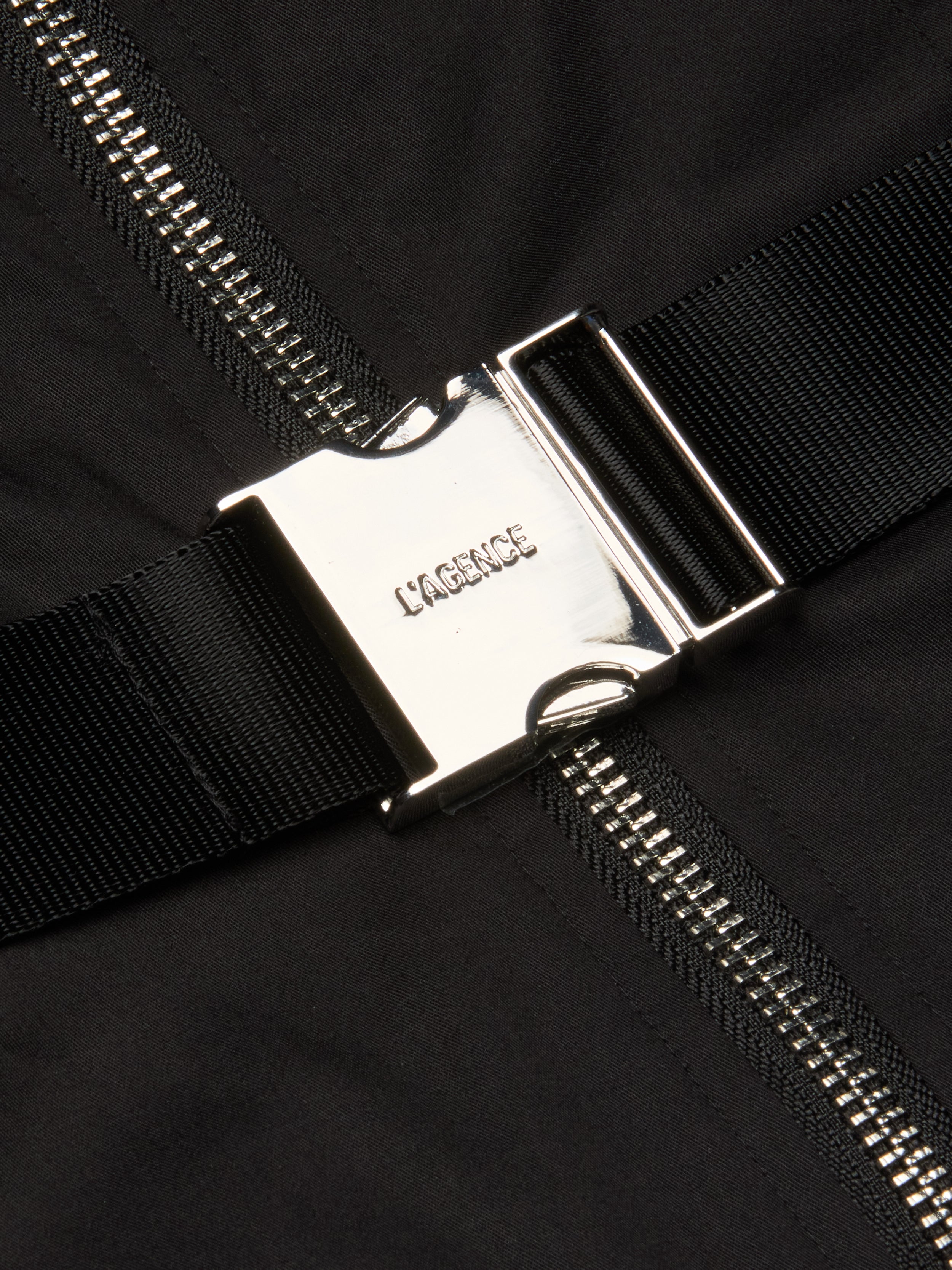 A close-up of a shiny silver buckle engraved with LAGENCE, fastened to a black strap on the Rima Maxi Dress in stretch poplin. Behind it, a metallic zipper runs vertically down the black fabric.