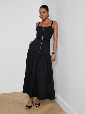 A woman stands against a white wall on wooden flooring wearing the sleeveless Rima Maxi Dress in black stretch poplin with a front zipper, wide belt, large earrings, bracelet, and black high-heeled sandals.
