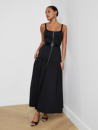 A woman stands against a plain white background wearing the Rima Maxi Dress— a sleeveless black maxi crafted from stretch poplin with a belted waist—paired with black sandals.