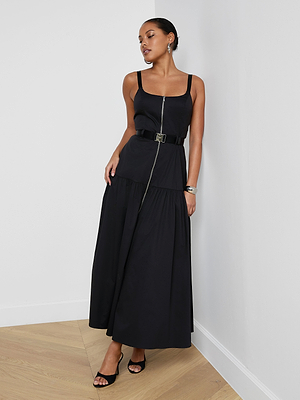 A woman stands indoors wearing the Rima Maxi Dress, a sleeveless black stretch poplin maxi with a square neck, front zipper, belted waist, and black open-toe heels. Her hair is pulled back and she accessorizes with earrings and a bracelet.