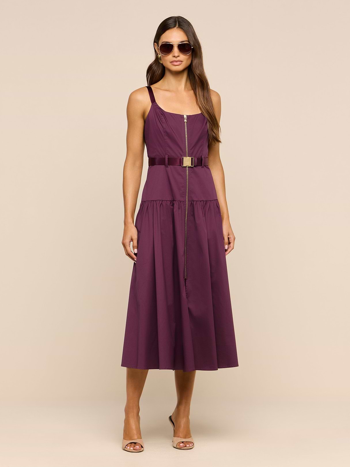 A woman wearing sunglasses and the Rima Dress—a sleeveless stretch poplin purple maxi dress with an adjustable belt—paired with strappy heels stands against a plain white background.