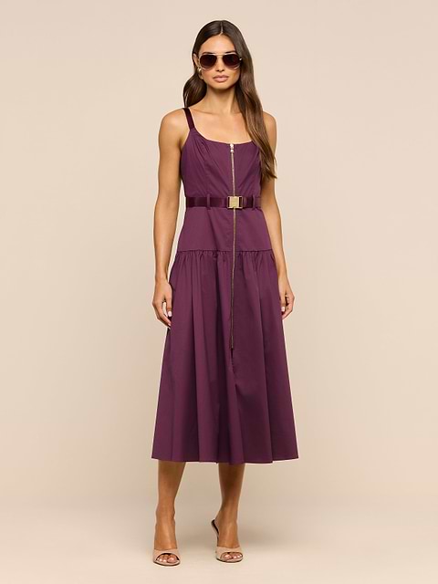 A woman wearing sunglasses and the Rima Dress—a sleeveless stretch poplin purple maxi dress with an adjustable belt—paired with strappy heels stands against a plain white background.