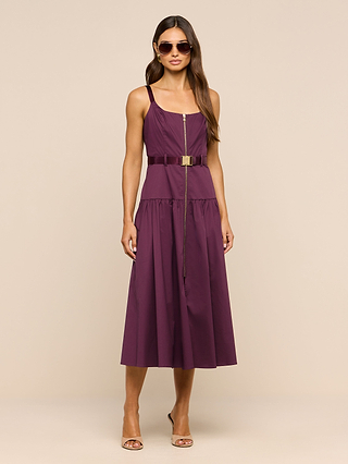 A woman stands against a plain beige background wearing sunglasses, open-toe heels, and the Rima Dress—a sleeveless, belted, purple stretch poplin midi dress with a front zipper.