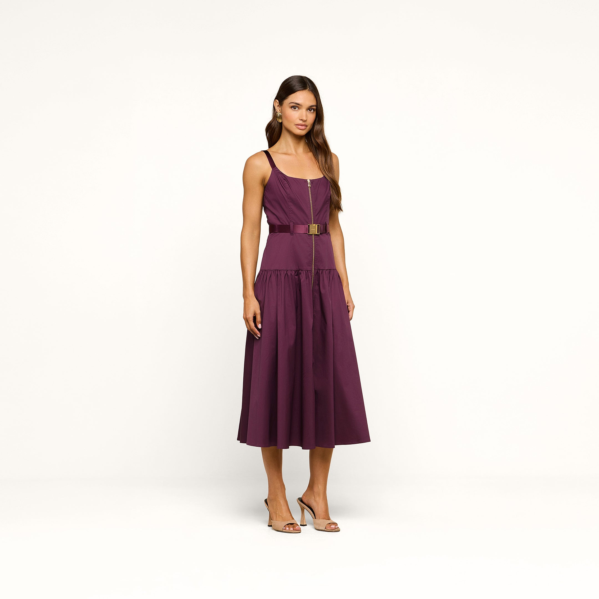 A woman stands against a plain white background wearing the Rima Dress, a sleeveless, belted purple stretch poplin midi dress, paired with high-heeled sandals.