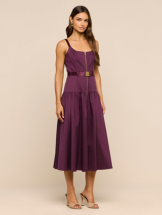 A woman stands in heels against a plain beige background, wearing the Rima Dress—a sleeveless, stretch poplin purple midi dress featuring a front zipper and an adjustable belt.