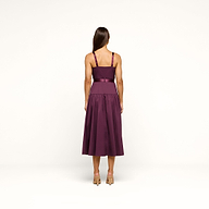 A woman stands facing away, modeling the Rima Dress—a sleeveless stretch poplin piece in deep purple with a fitted bodice, wide waistband, and a calf-length flared skirt. She has long brown hair and is wearing nude heels against a plain white background.