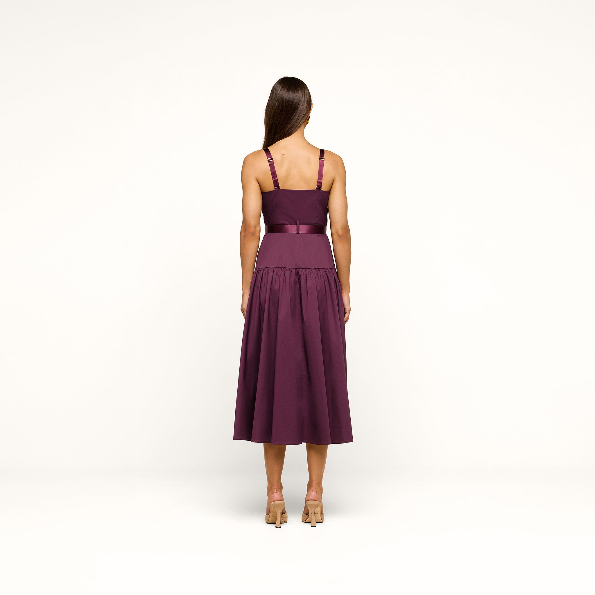A woman stands facing away, modeling the Rima Dress—a sleeveless stretch poplin piece in deep purple with a fitted bodice, wide waistband, and a calf-length flared skirt. She has long brown hair and is wearing nude heels against a plain white background.