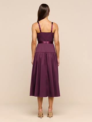 A woman stands facing away, wearing the Rima Dress—a sleeveless, dark purple stretch poplin midi dress with a fitted bodice and flared skirt. She has long, straight brown hair and beige high-heeled sandals. The background is plain beige.