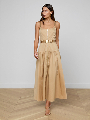 A woman with long brown hair and gold earrings stands indoors on a light wood floor wearing the Rima Maxi Dress—a sleeveless, neutral tan stretch poplin maxi with a square neckline, belted waist, and strappy sandals against a plain wall.