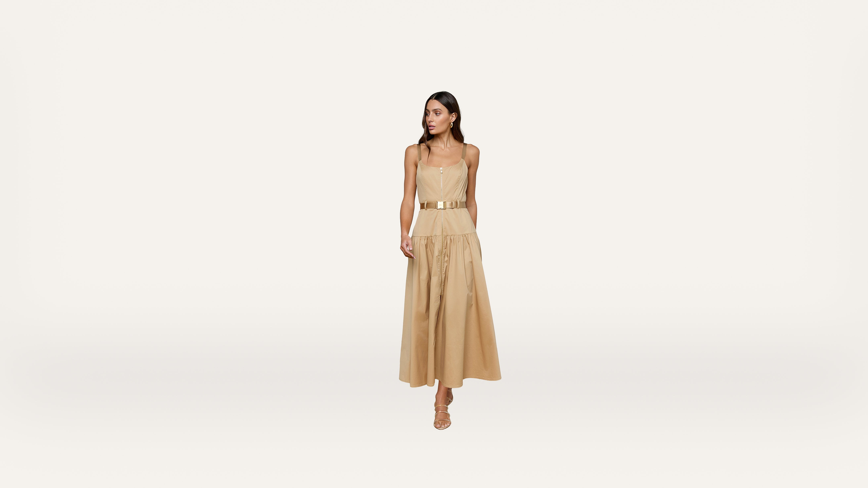 A woman wearing a tan sleeveless belted midi dress and strappy sandals stands against a plain white background.