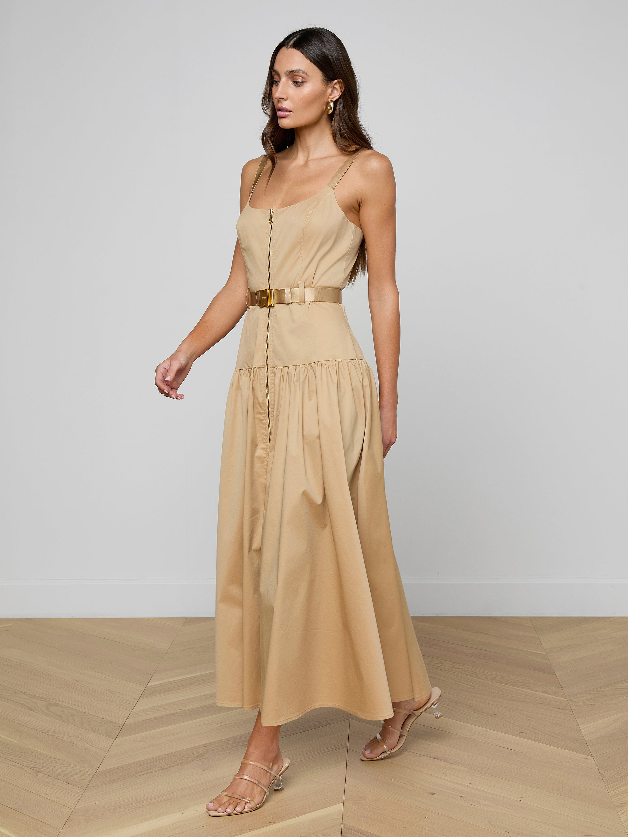 A woman wearing the Rima Maxi Dress—a neutral tan, stretch poplin sleeveless dress with a belted waist—walks in strappy heels against a plain white background.