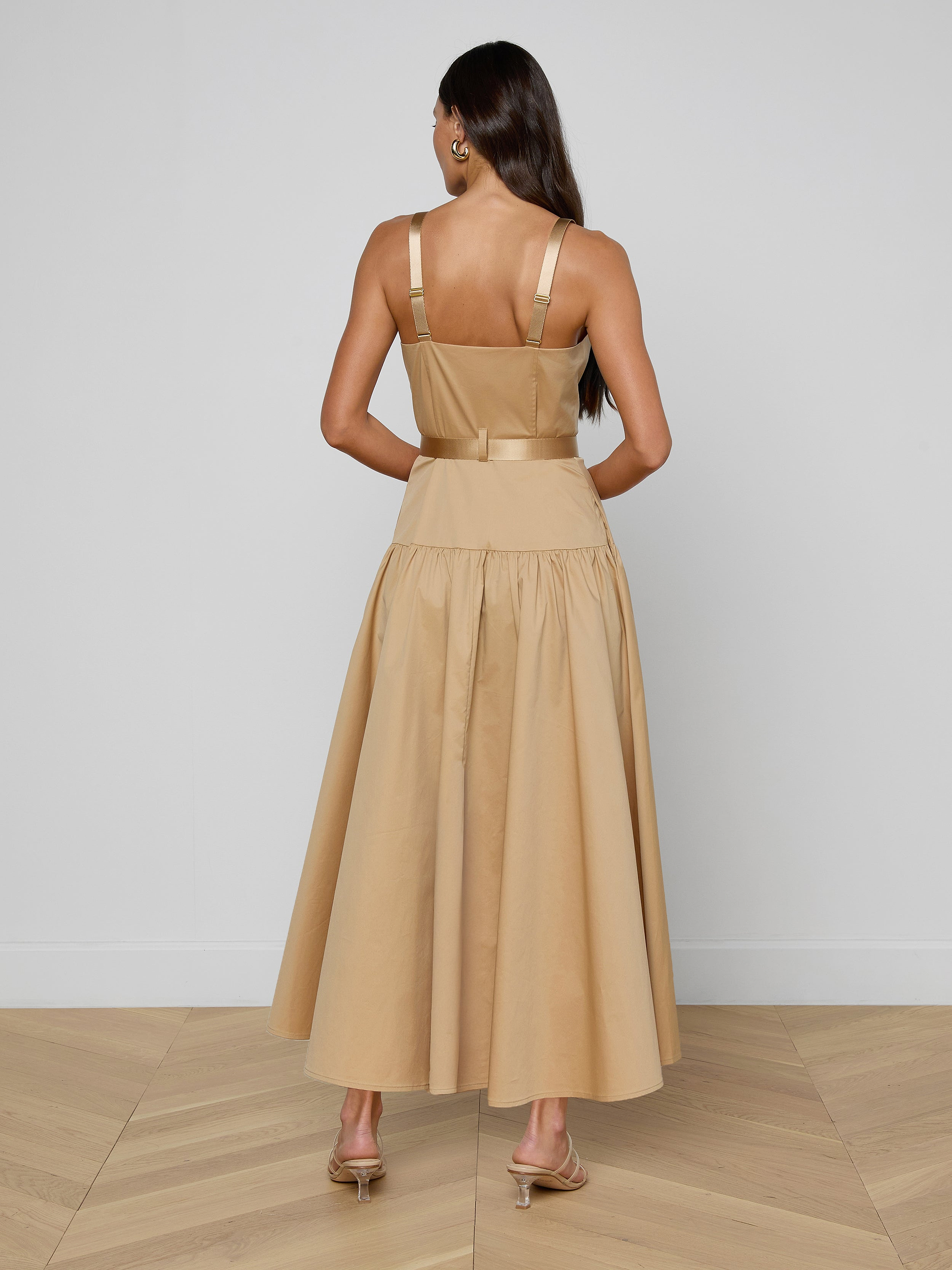 A woman with long dark hair is wearing the Rima Maxi Dress in neutral tan stretch poplin and heels, standing with her back to the camera against a plain white background.