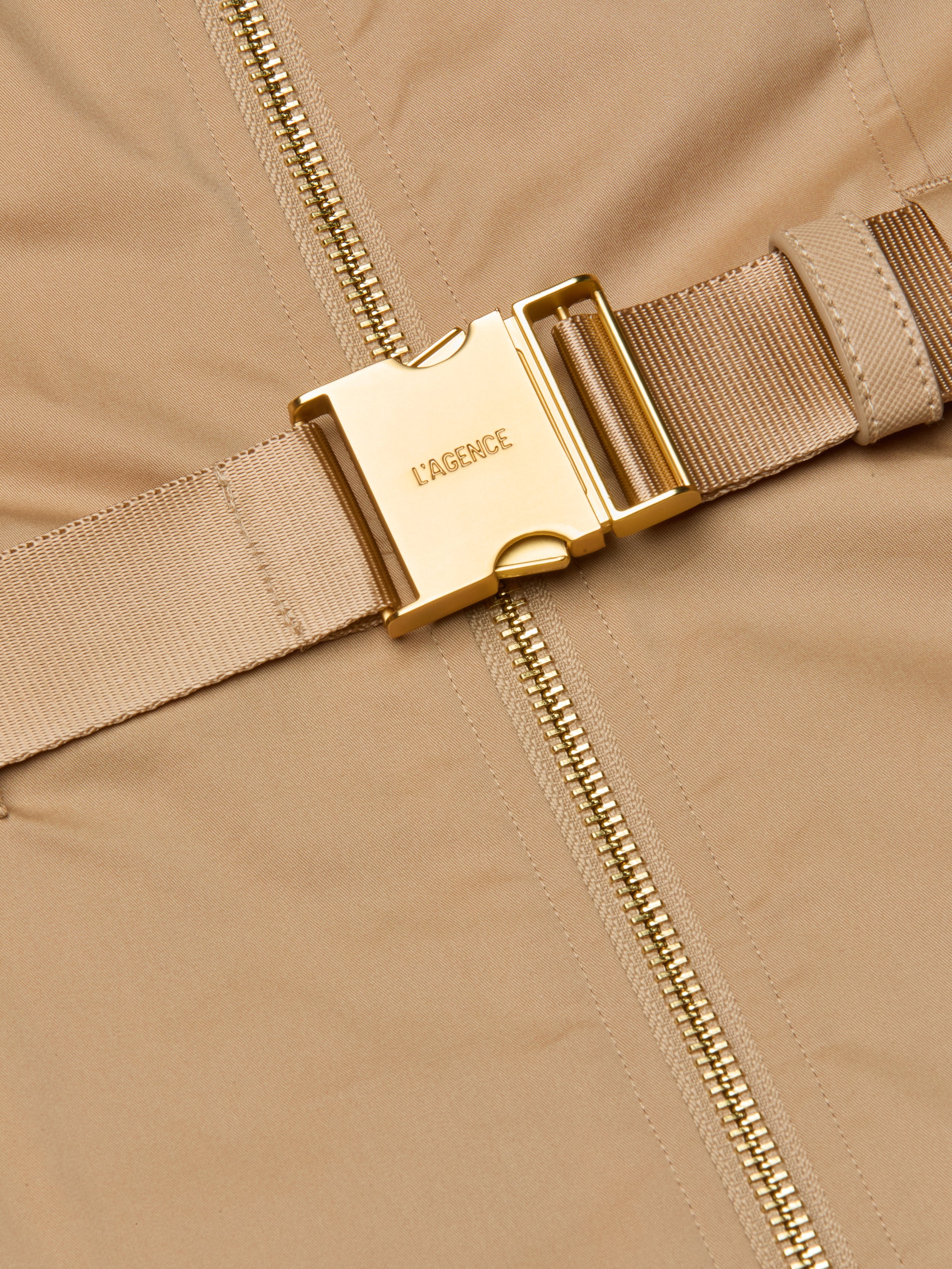 Close-up of the Rima Maxi Dress in a neutral tan stretch poplin, showcasing a gold metal buckle engraved with LAGENCE and a beige zipper running vertically beside it. The buckle is attached to a matching fabric strap.