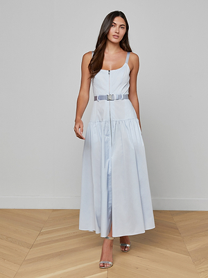 A woman with long brown hair stands indoors on a wooden floor, wearing the pale blue Rima Maxi Dress with a front zipper and matching belt, paired with silver open-toe heels against a plain light-colored wall.
