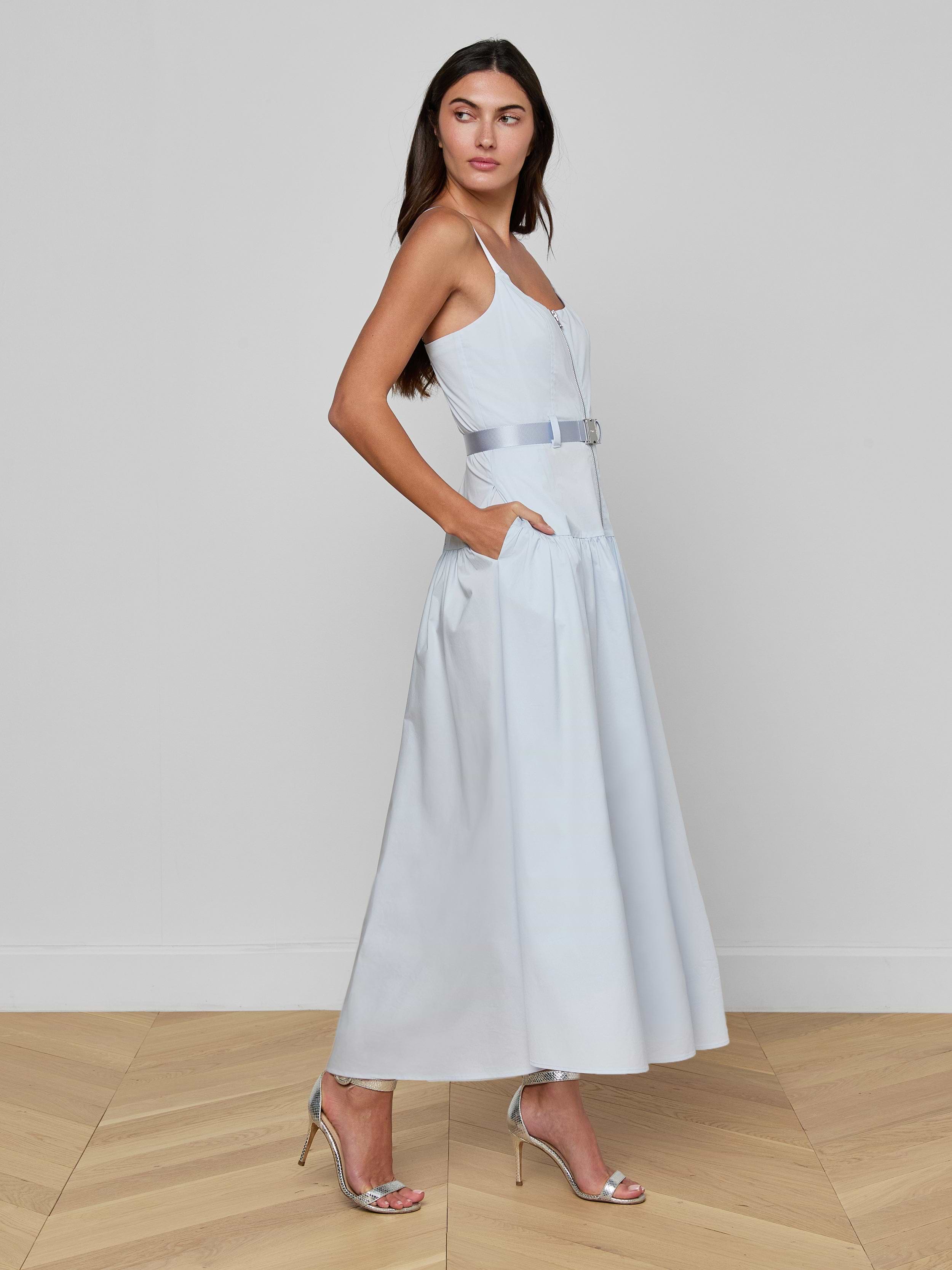A woman stands sideways against a plain white background wearing the Rima Maxi Dress, a pale blue stretch poplin maxi style featuring a belt and pockets, paired with silver high-heeled sandals.