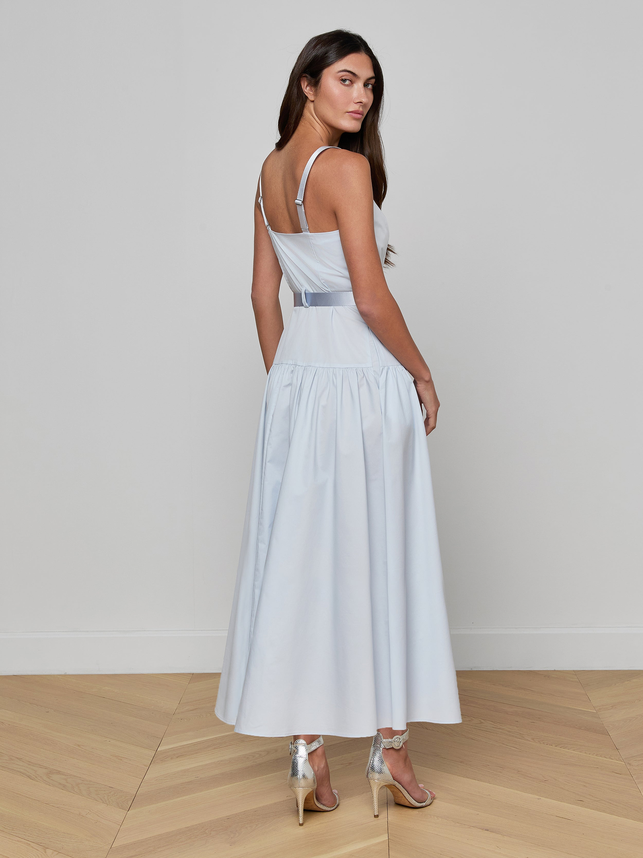 A woman with long dark hair stands against a plain light background, wearing the Rima Maxi Dress—a sleeveless pale blue stretch poplin maxi—and high-heeled sandals, looking over her shoulder.