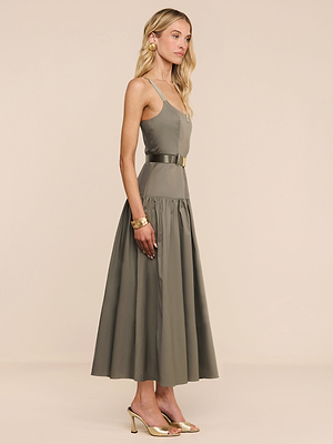 A woman stands in profile against a neutral background, wearing the Rima Dress—an olive green dress with adjustable straps and a belted waist—paired with gold heels, pink earrings, and gold bracelets.