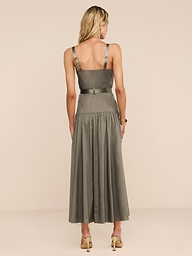A woman with blonde hair faces away, wearing the Rima Dress—an olive green maxi with a fitted bodice, flared skirt, and adjustable straps—paired with beige high-heeled sandals against a plain beige background.